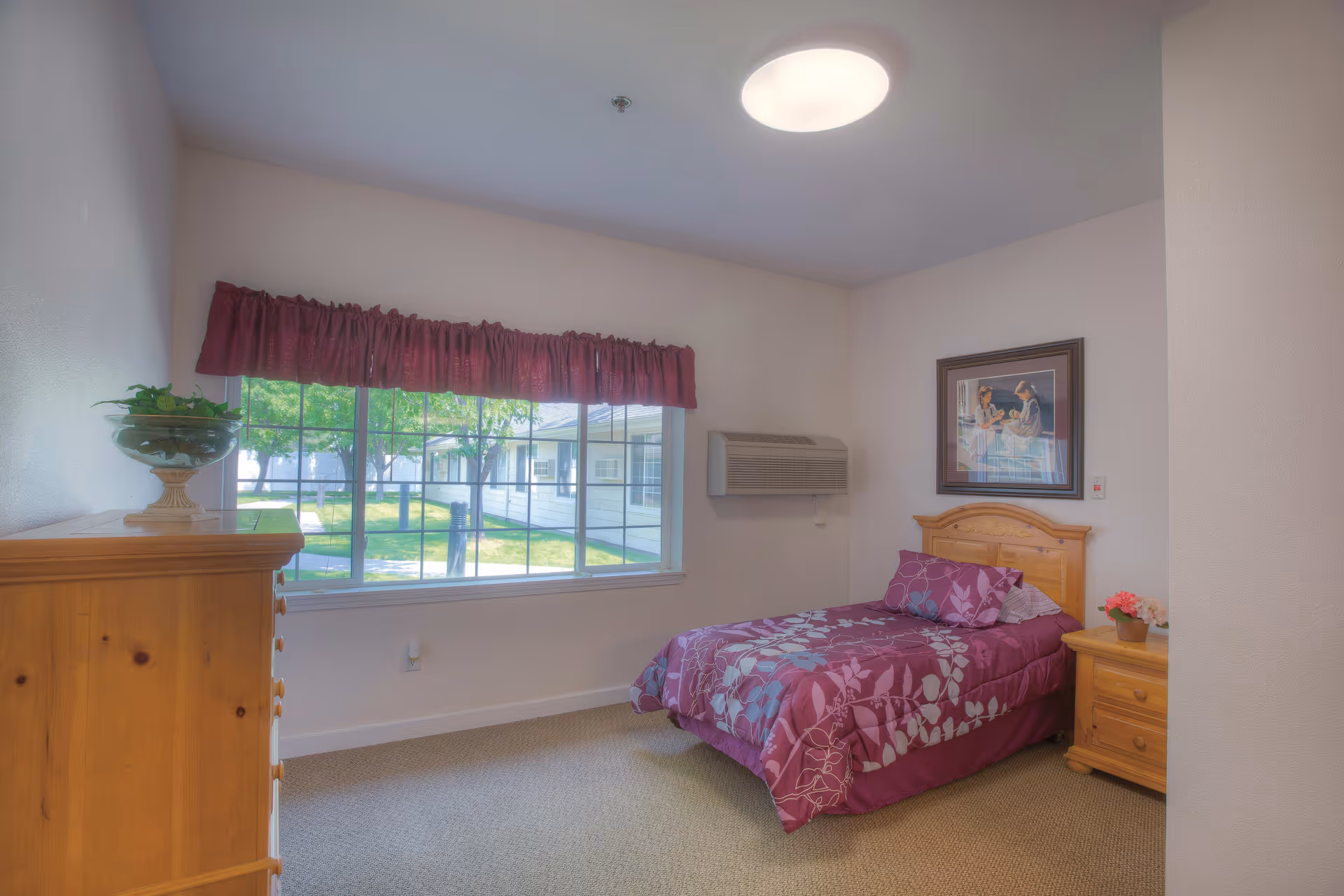 A small, cozy bedroom with a single bed covered in a purple floral bedspread. There is a wooden nightstand with a small flower pot and a wooden dresser with a glass bowl on top. A large window with a red valance shows a view of green trees and a building outside. A framed picture hangs on the wall above the bed, and an air conditioning unit is mounted on the wall.