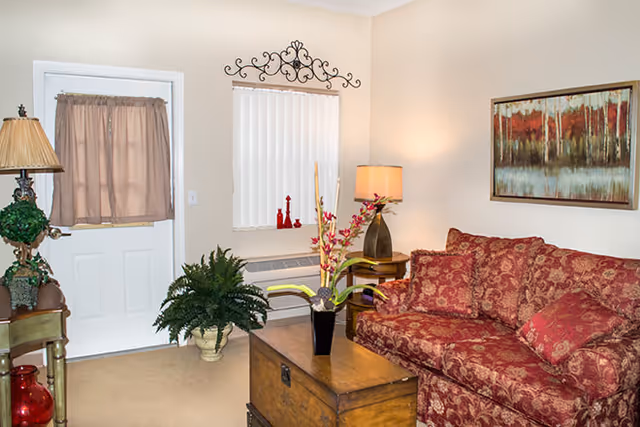 Small living room with a patterned red sofa, wooden trunk coffee table, potted plants, and a lamp beside a door and window.