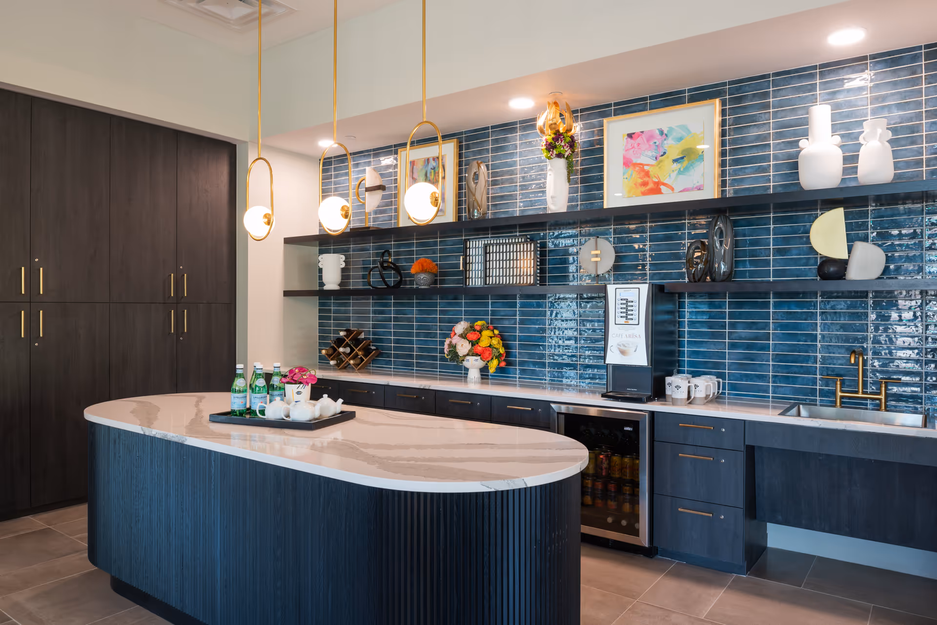 Modern kitchen area with a large curved island countertop, three pendant lights hanging above, dark wood cabinets, blue tiled backsplash, decorative shelves with art pieces and flowers, a small beverage refrigerator, and a sink with a gold faucet.