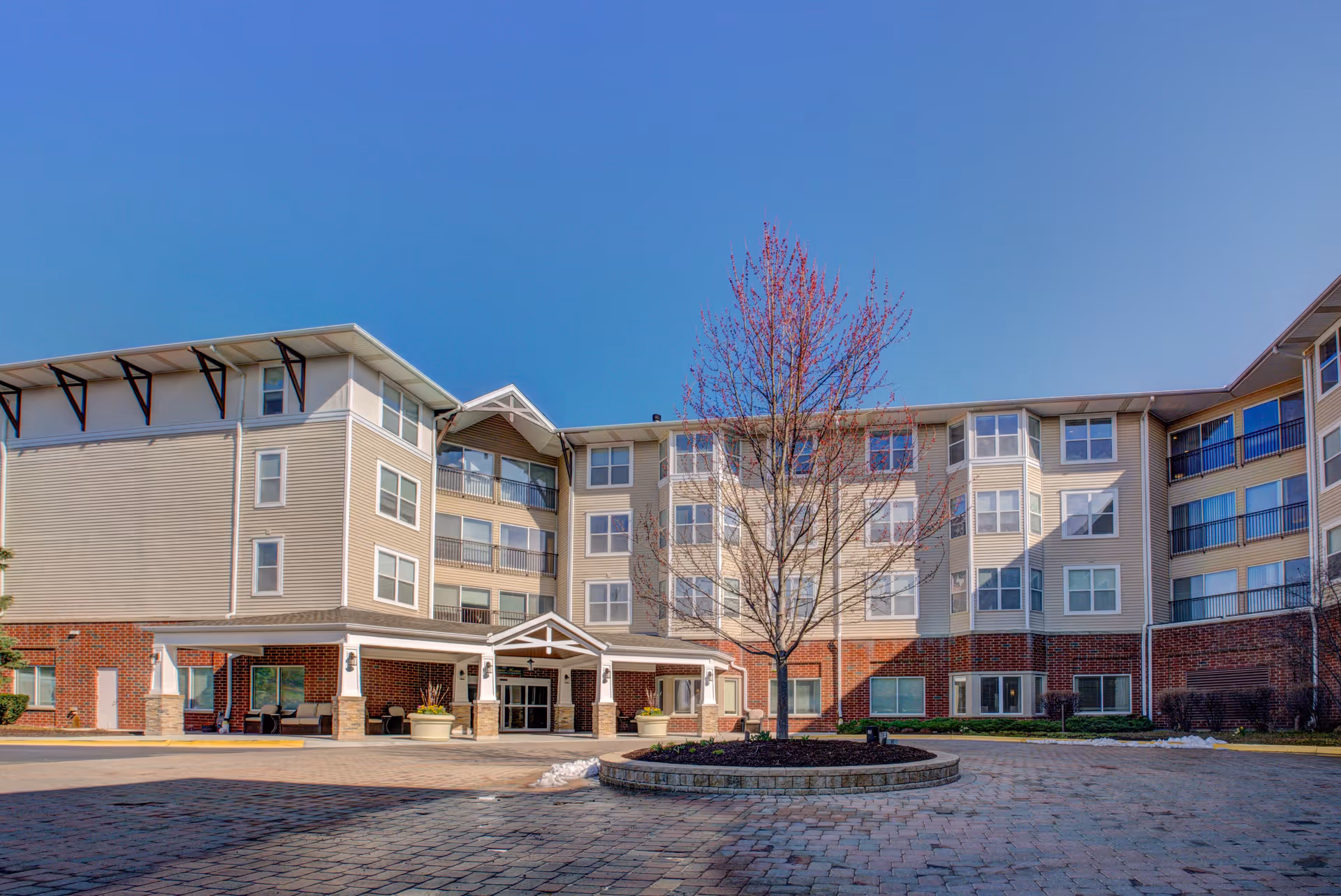 Exterior view of a multi-story senior living facility building with a covered entrance, large windows, and a tree in the center of a circular landscaped area in front of the building under a clear blue sky.
