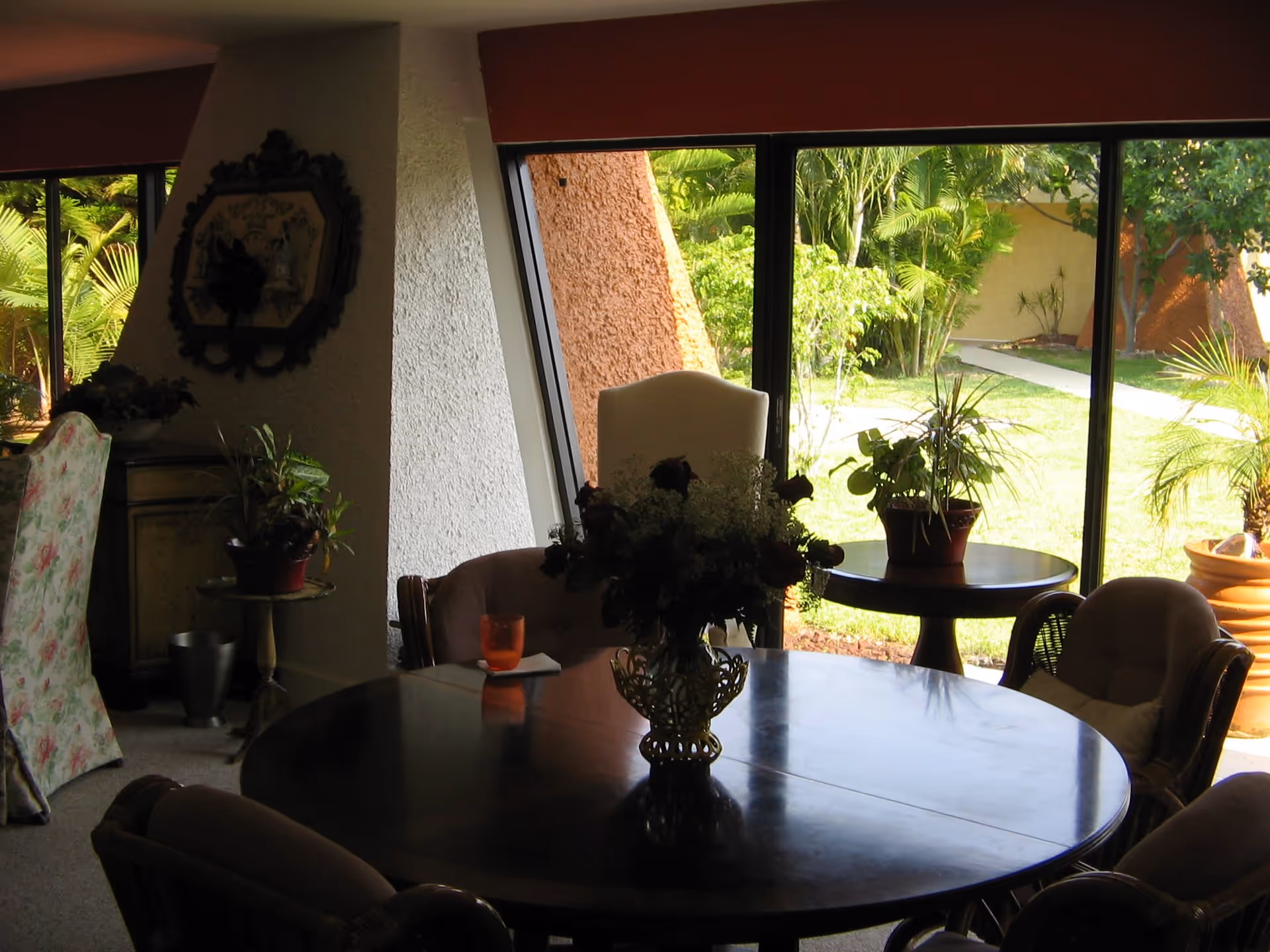 Interior view of a living room or common area with a round wooden table in the center, surrounded by cushioned chairs. A vase with flowers is placed on the table. Large windows in the background reveal a green outdoor garden with plants and a walkway. Additional potted plants and furniture are visible near the windows.