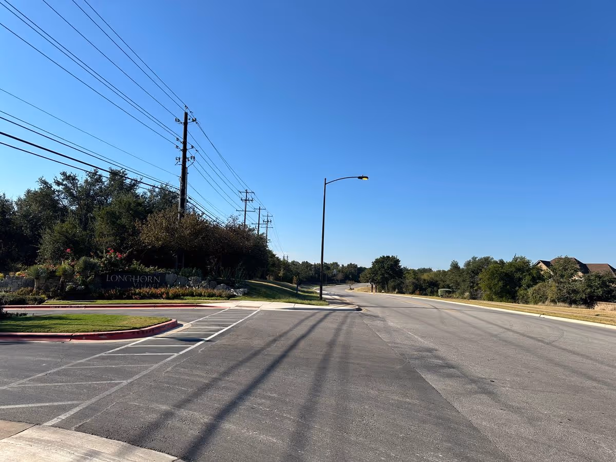 Wide empty roadway and entrance with a streetlight, utility poles and a landscaped stone sign reading 'LONGHORN' under a clear blue sky.