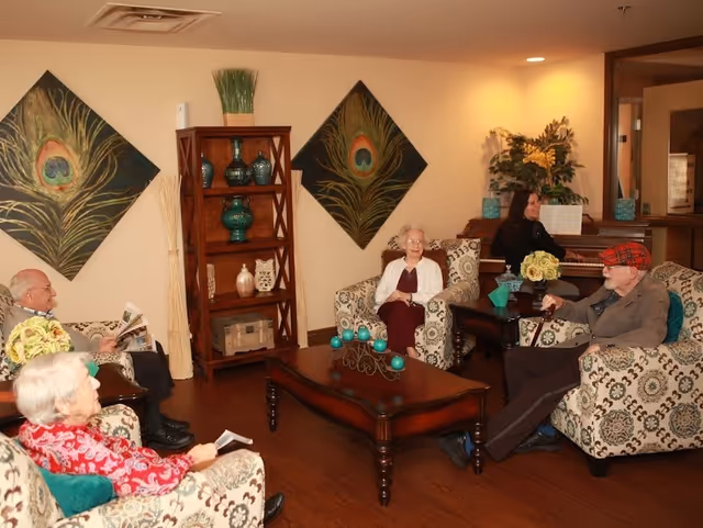 A cozy living room in a senior living facility with four elderly people seated on patterned armchairs around a wooden coffee table decorated with teal ornaments. A woman is playing the piano in the background. The walls are decorated with two large peacock feather paintings and a wooden shelf holding vases and decorative items.