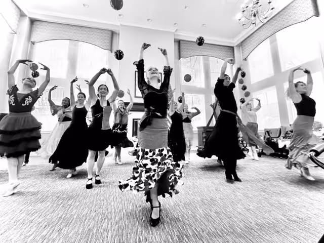 A group of dancers performing indoors in a spacious room with large windows and high ceilings. The dancers are dressed in various costumes and are captured mid-movement with their arms raised.