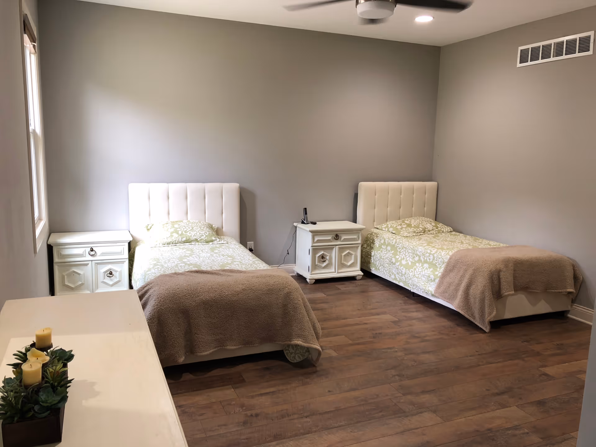 A simple bedroom with two single beds, each with a white upholstered headboard and green patterned bedding with beige blankets. There are two white nightstands between and beside the beds. The room has wooden flooring, gray walls, a window on the left, and a ceiling fan with a light.