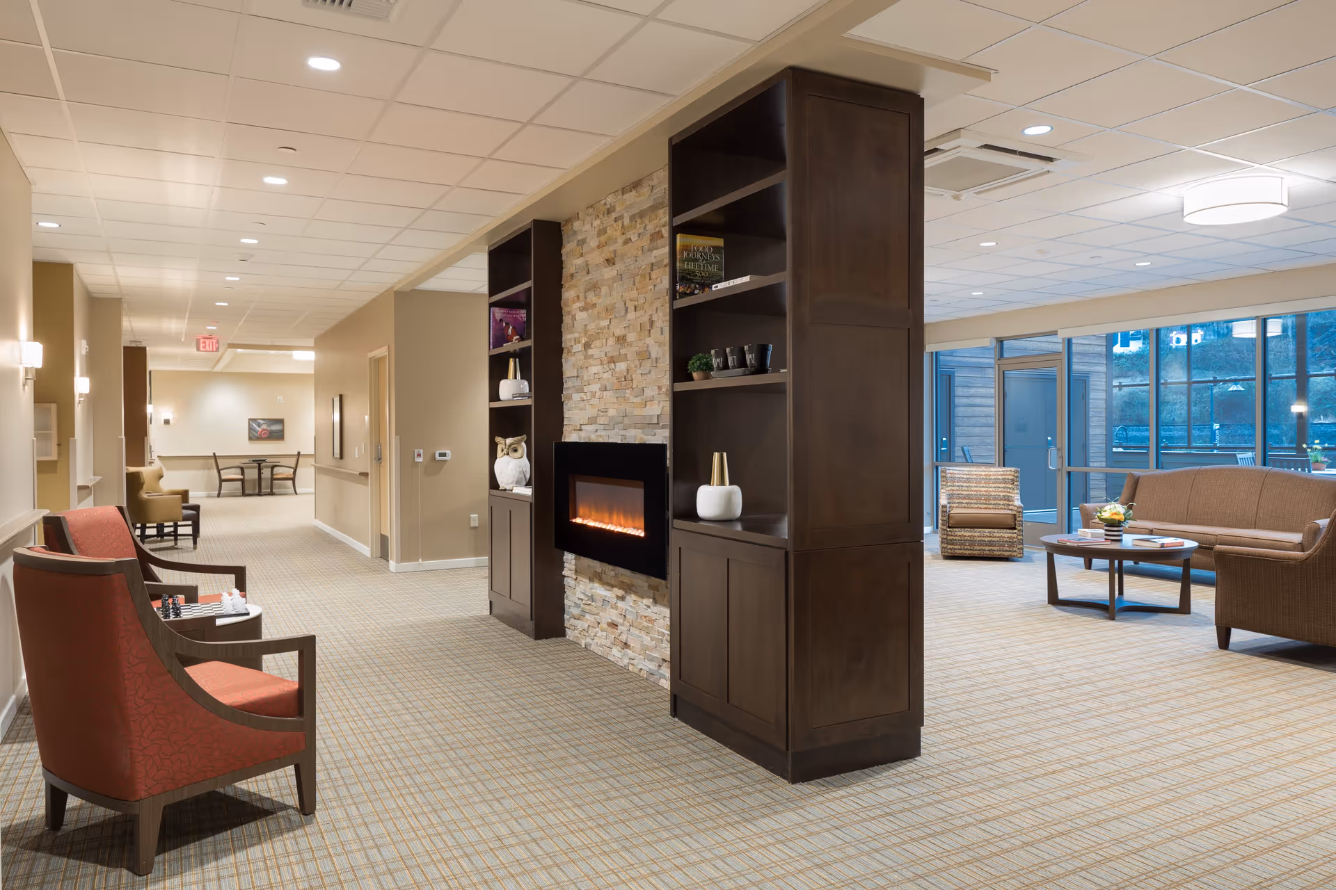 A spacious and well-lit interior common area in a senior living facility featuring a modern electric fireplace set in a stone wall with dark wooden shelving units on either side. The room includes comfortable seating such as red armchairs with a chess set on a small table, a brown sofa, and a patterned armchair near large windows that let in natural light. The carpet has a subtle plaid pattern, and the ceiling has recessed lighting and a round ceiling light fixture.
