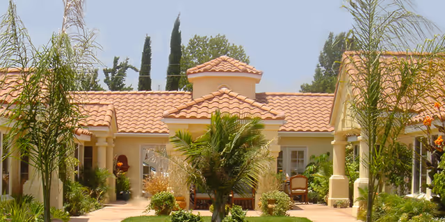 Exterior view of a single-story building with a terracotta tiled roof, surrounded by palm trees and other greenery under a clear sky.