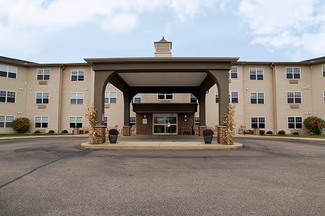 Front exterior view of Dimensions Living Stevens Point building with a covered entrance and a paved driveway in front. The building is three stories tall with multiple windows and beige siding.