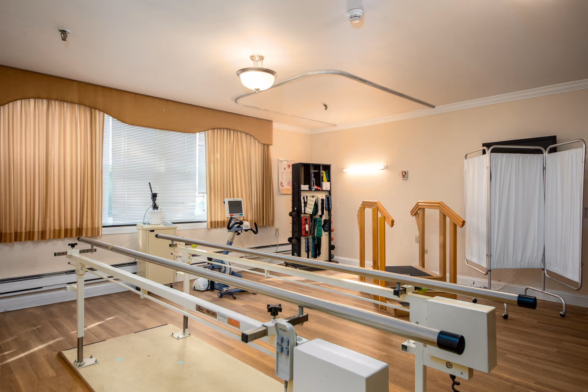 A rehabilitation or physical therapy room with parallel bars for walking exercises, a stationary exercise bike, a small set of wooden stairs with handrails, a privacy screen, and a storage rack with various therapy equipment. The room has wood flooring, beige walls, a window with closed blinds and beige curtains, and ceiling lighting.