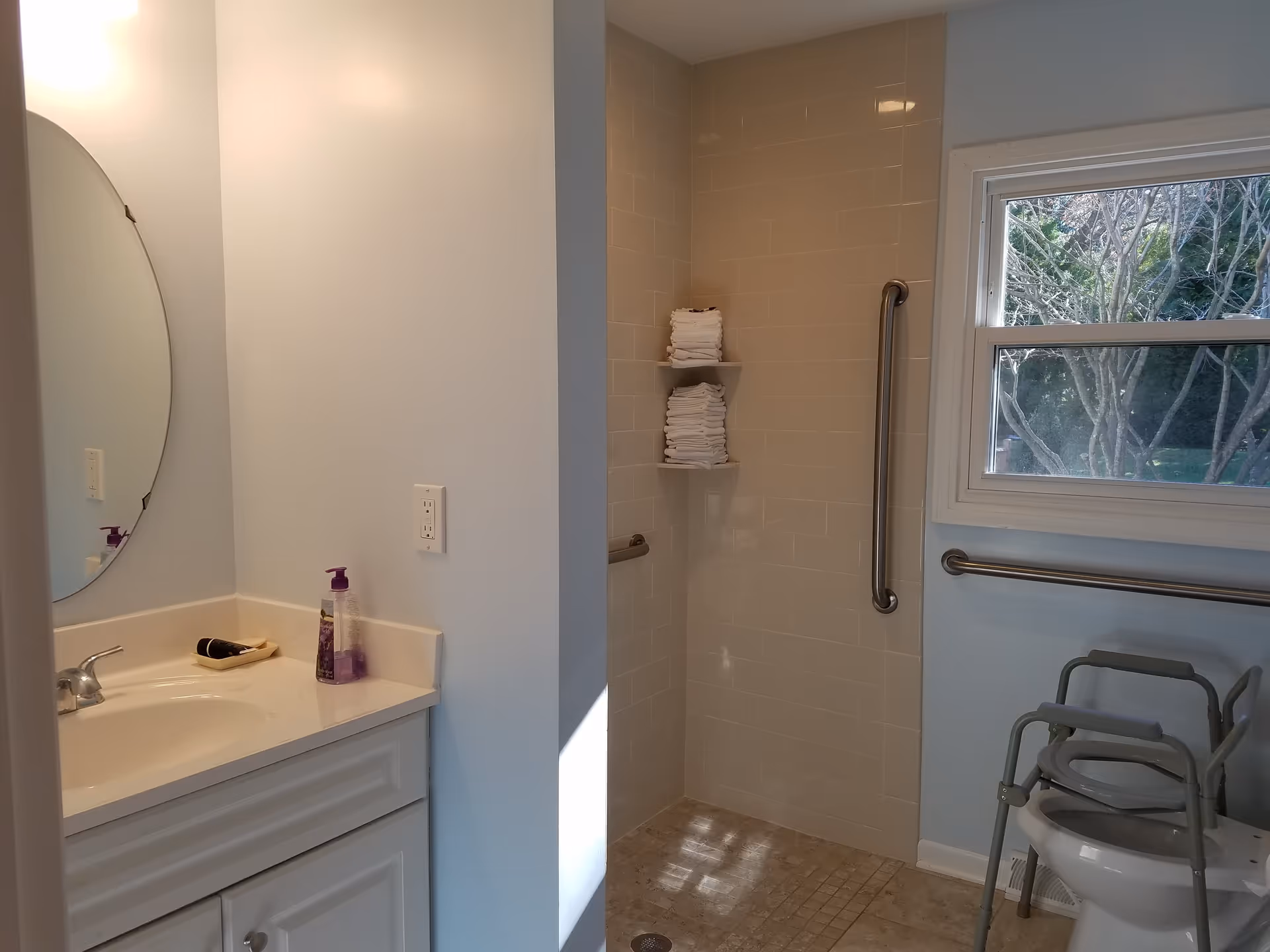 A bathroom with a white vanity and sink on the left, a round mirror above the sink, a walk-in tiled shower with two shelves holding folded white towels, grab bars on the shower wall and near the window, and a toilet with a raised seat and armrests on the right side under a window showing trees outside.