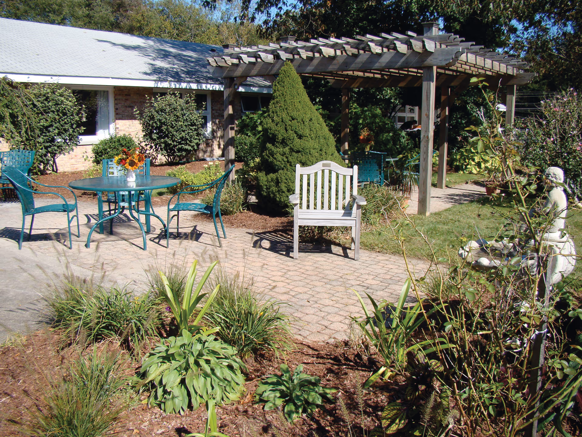 Outdoor garden area with a paved patio featuring a round green metal table with four matching chairs, a wooden bench, and a wooden pergola. There are various plants, shrubs, and a small statue in the garden surrounding the patio. A building with brick walls and white-framed windows is visible in the background.