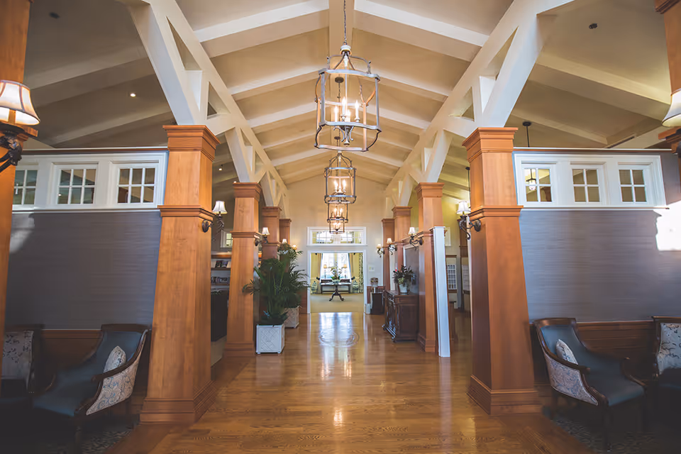 Spacious interior hallway with wooden floors, large wooden columns, and vaulted ceiling with exposed beams. Hanging lantern-style light fixtures illuminate the space. Seating areas with upholstered chairs are visible on both sides, along with potted plants and wall-mounted lamps.