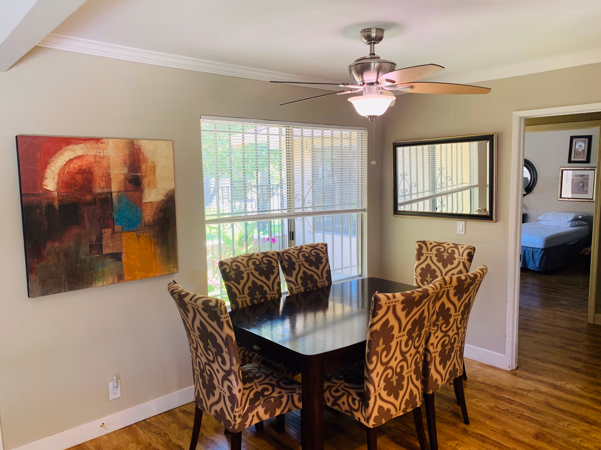 A dining area with a dark wooden table surrounded by six upholstered chairs with a brown and beige patterned fabric. A large window with blinds lets in natural light, and a ceiling fan with a light fixture is above the table. On the left wall, there is an abstract painting with warm colors, and on the right wall, a large mirror reflects part of the room. Through an open doorway, a bedroom with a bed and framed pictures on the wall is visible.