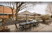 Outdoor patio area with a rectangular table and eight chairs under two large beige umbrellas. Surrounding the patio are trees and shrubs with a brick building visible in the background under a cloudy sky.