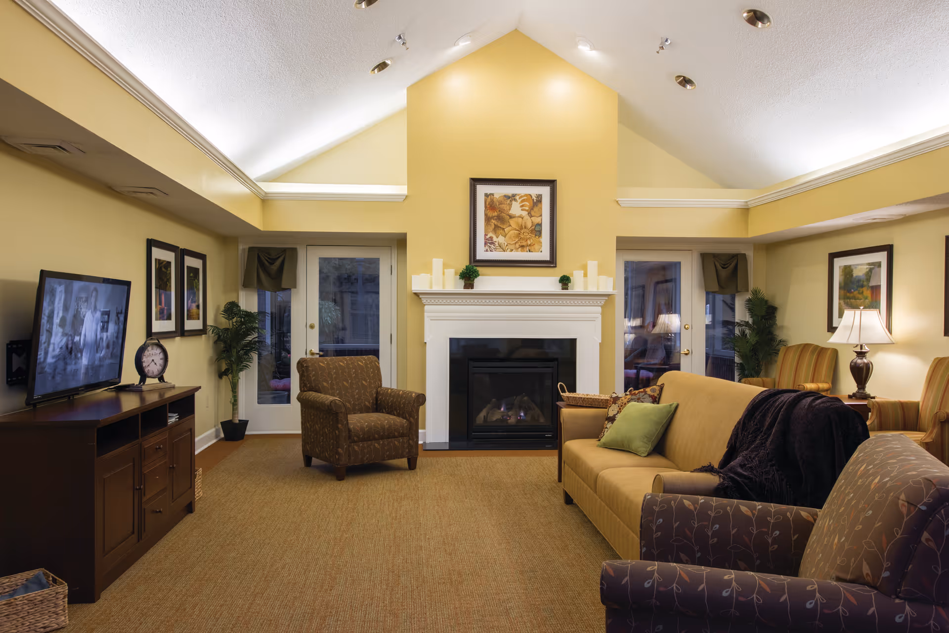 A cozy living room with a high vaulted ceiling painted in light yellow. The room features a white fireplace with candles and a framed floral painting above it. There are two glass doors on either side of the fireplace with plants nearby. The seating includes a brown patterned armchair, a mustard yellow sofa with green and patterned pillows, and another armchair with a floral pattern. A TV is mounted on a wooden cabinet on the left side, and there are framed pictures on the walls. The room is warmly lit with lamps and recessed ceiling lights.