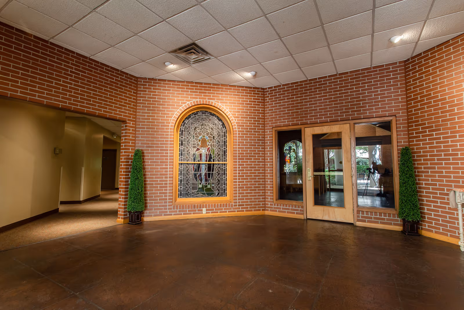 Indoor lobby area with brick walls, a stained-glass window, double glass doors, and tall potted topiary plants.
