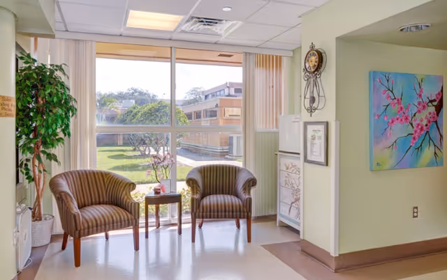 A bright sitting area with two striped armchairs and a small wooden table between them, placed in front of large windows showing a green outdoor courtyard. The room has light-colored walls, a potted plant on the left, a decorative wall clock, a framed certificate, and a colorful painting of pink flowers on the right wall.