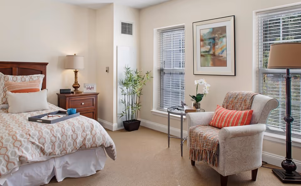 A cozy bedroom with a neatly made bed featuring patterned bedding and multiple pillows. Next to the bed is a wooden nightstand with a lamp, a clock, and a framed photo. There is a potted plant in the corner near two windows with blinds. An upholstered armchair with a striped pillow and a throw blanket sits beside a small round side table holding a white orchid and a book. A floor lamp stands near the windows, and a framed abstract painting hangs on the wall.