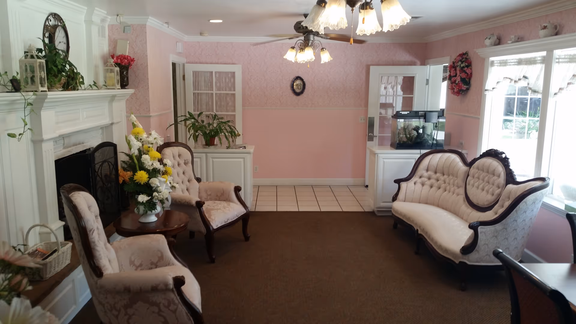 A cozy living room with vintage-style upholstered chairs and a sofa arranged around a small wooden table with a flower arrangement. The room features pink wallpaper, a white fireplace with plants and lanterns on the mantel, a ceiling fan with lights, a fish tank on a cabinet, and large windows with lace curtains letting in natural light.