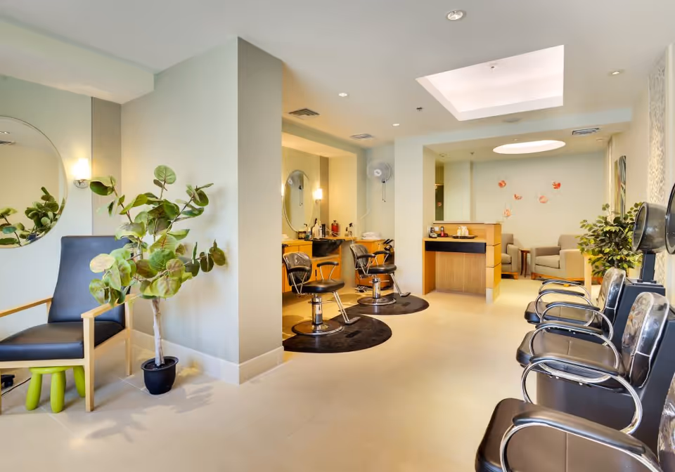 Interior view of a bright and modern hair salon area in a senior living facility. The room features black salon chairs with mirrors and hair washing stations, a waiting area with black chairs, potted plants, and soft lighting from ceiling fixtures and wall sconces.