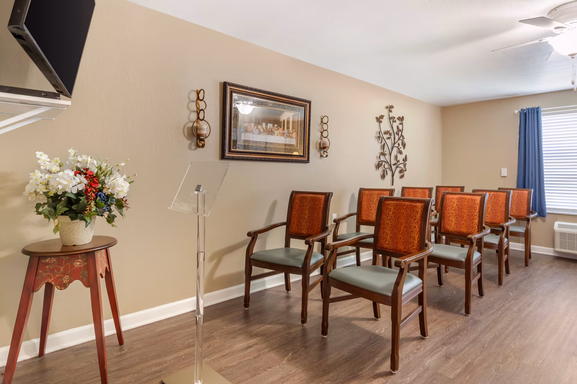 A small meeting or presentation room with eight wooden chairs with red patterned backs and green seats arranged in two rows. A clear acrylic lectern stands at the front left of the chairs. On the left side, there is a small wooden table with a floral arrangement in a vase. The beige walls are decorated with a framed picture, two candle holders, and a metal wall art piece shaped like a tree. A window with blue curtains and a ceiling fan with a light are visible on the right side.