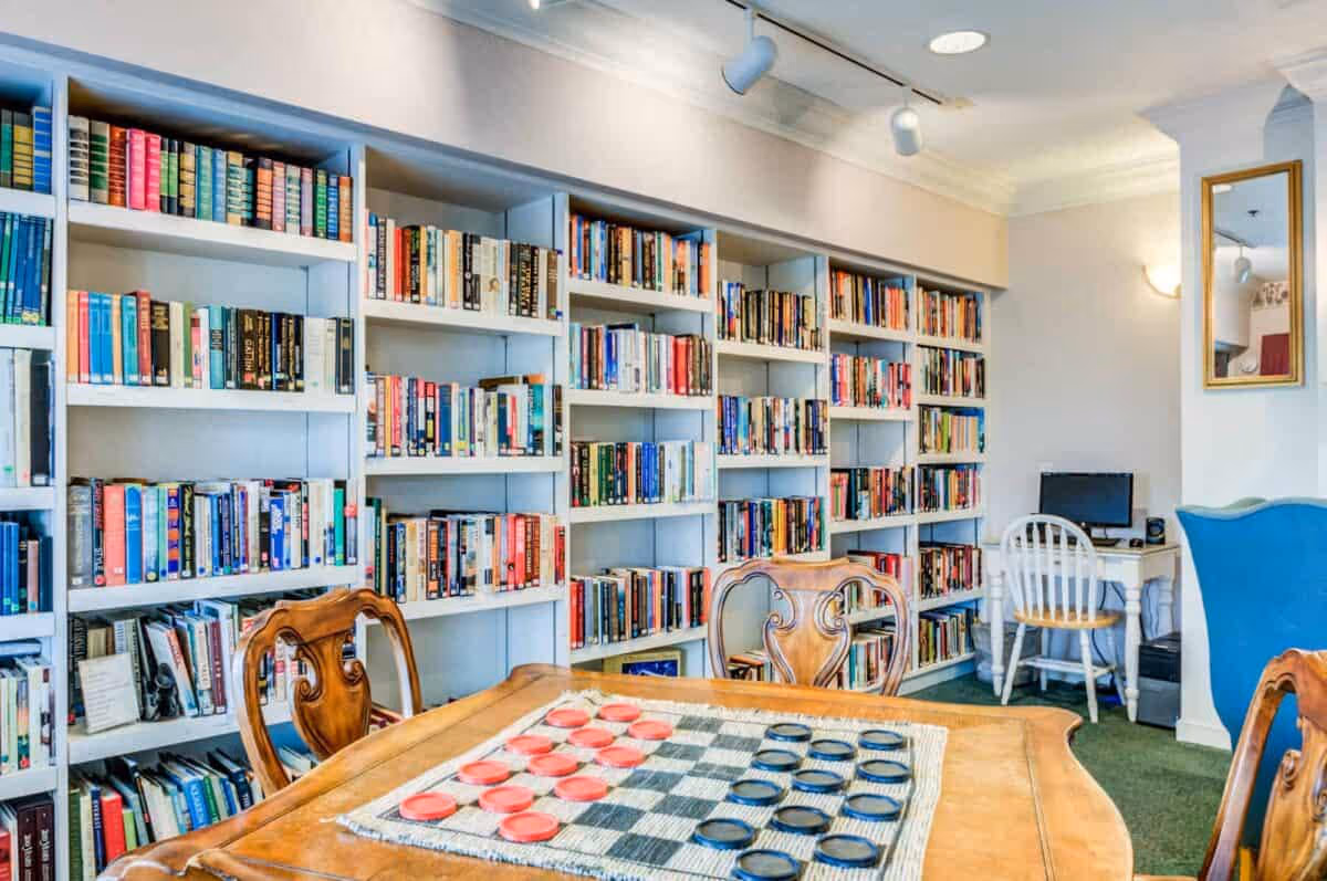 Cozy interior reading room with wall-to-wall bookshelves, a table set up for checkers, several chairs, and a small computer desk in the corner.
