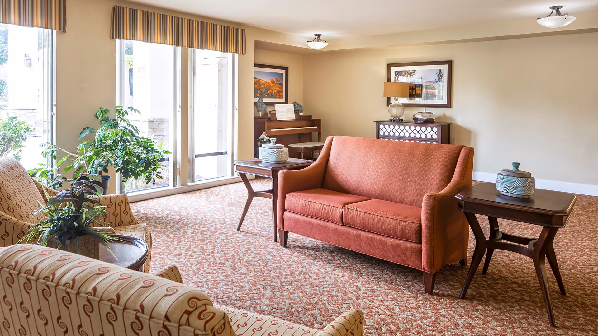 A cozy living room area with a red upholstered sofa, two patterned armchairs, wooden side tables with decorative items, a piano in the corner, and large windows with striped valances letting in natural light. The room has a patterned carpet and neutral-colored walls with framed artwork.