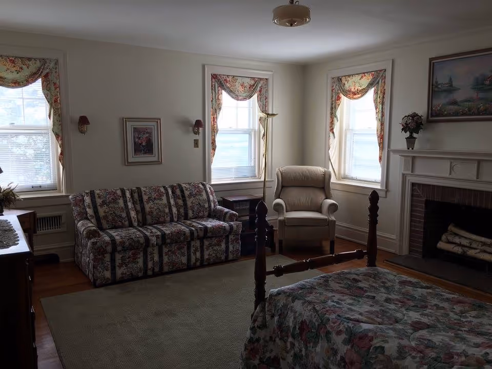 A cozy room with a floral patterned bedspread on a wooden bed frame, a matching floral sofa, and a beige armchair. The room has three windows with floral curtains, a fireplace with logs, a painting above the mantel, and a floor lamp next to the armchair. The walls are light-colored and the floor has a large area rug.