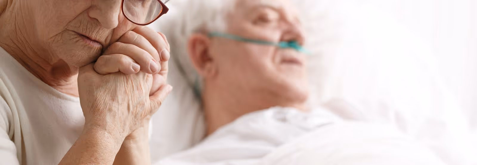 An elderly woman with glasses is holding the hand of an elderly man lying in a hospital bed with an oxygen tube in his nose, showing a moment of care and support.