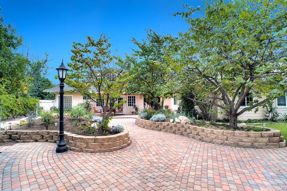 A paved courtyard with a brick pattern surrounded by raised garden beds containing trees and shrubs. A black lamppost stands on the left side. In the background, there is a single-story building with a porch and outdoor seating under a clear blue sky.