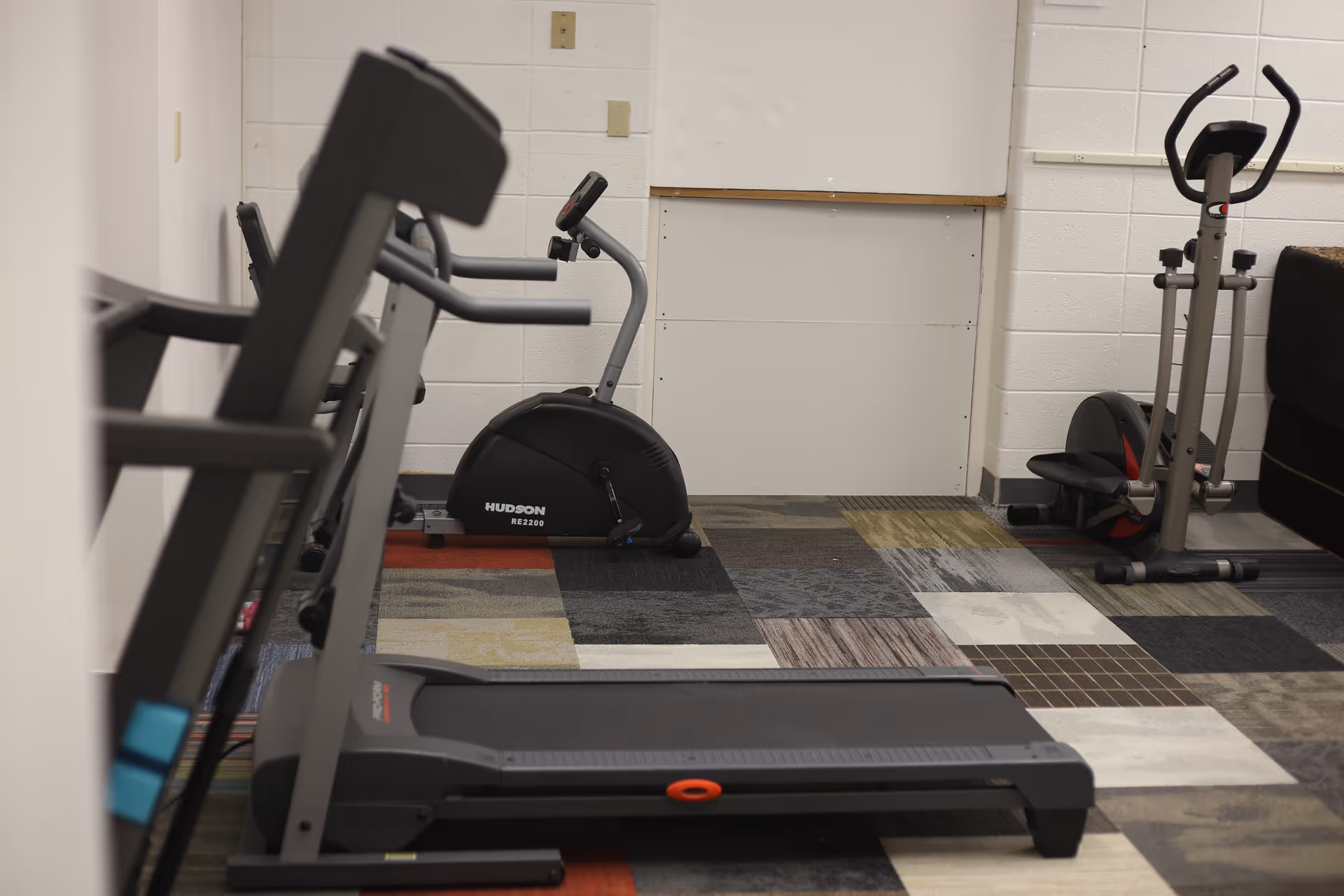 Indoor exercise room with a treadmill, a stationary bike, and an elliptical machine on a multicolored patterned carpet. The walls are white with a whiteboard mounted on one wall.