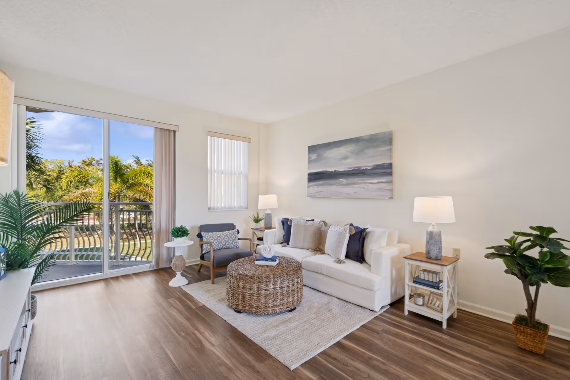 A bright and airy living room with a white sofa adorned with multiple cushions, a round woven coffee table on a light rug, a gray armchair with a patterned pillow, two side tables each with a lamp, and large sliding glass doors opening to a balcony with a view of palm trees and greenery.