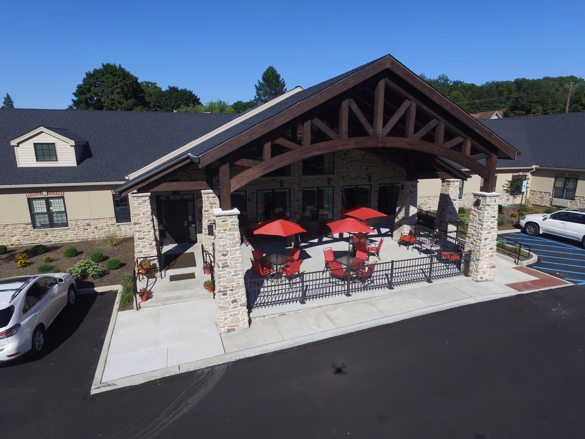 Exterior view of a senior living facility with a stone and beige facade, featuring a covered patio area with red umbrellas and matching red chairs. There are two cars parked in the adjacent parking lot, and the sky is clear and blue.