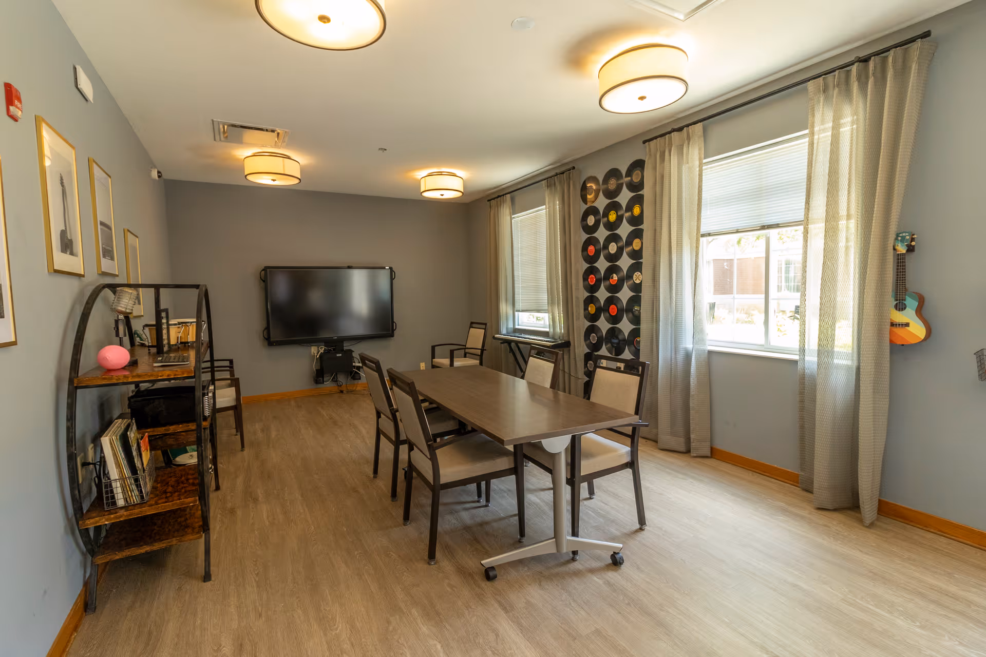 A well-lit room with a rectangular table surrounded by six chairs. The room has light wood flooring, gray walls, and two large windows with sheer curtains. On one wall, there is a collection of vinyl records displayed decoratively. A flat-screen TV is mounted on the opposite wall, and a shelving unit with books and decorative items is positioned nearby. A small colorful guitar hangs on the wall near the windows.