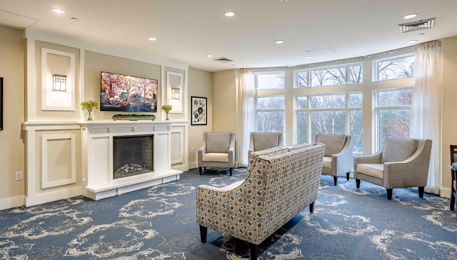 A bright and spacious living room area in a senior living facility with a patterned blue carpet, a white fireplace with a mounted flat-screen TV above it, and several upholstered chairs and a sofa arranged around the room. Large windows with sheer curtains allow natural light to fill the space, and decorative wall sconces and framed artwork add to the cozy ambiance.
