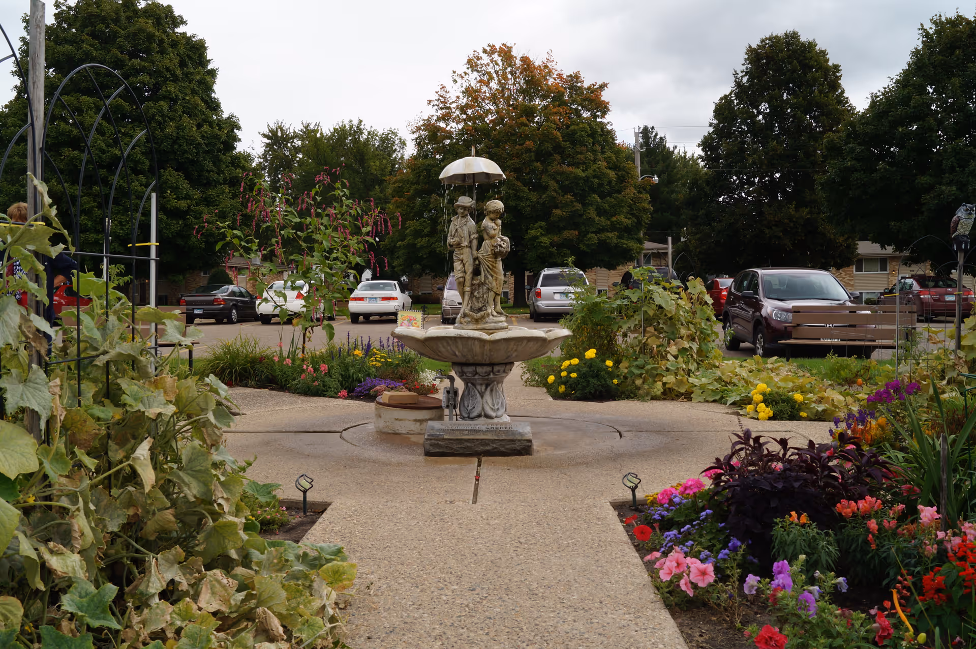A garden area with a central decorative fountain featuring a statue of two children under an umbrella. The garden is surrounded by colorful flowers and greenery, with a paved walkway leading to the fountain. Several parked cars and trees are visible in the background under a cloudy sky.