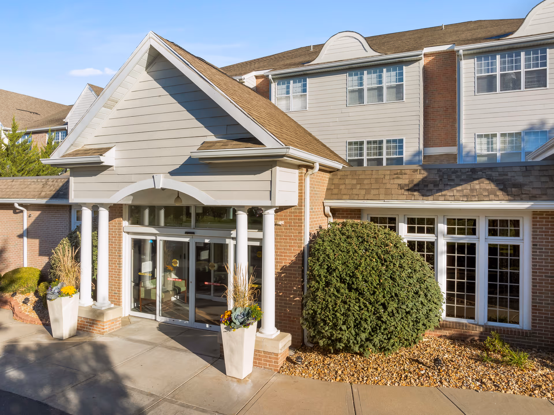 Front entrance of a multi-story senior living building with white columns, glass automatic doors, planters, and large windows.