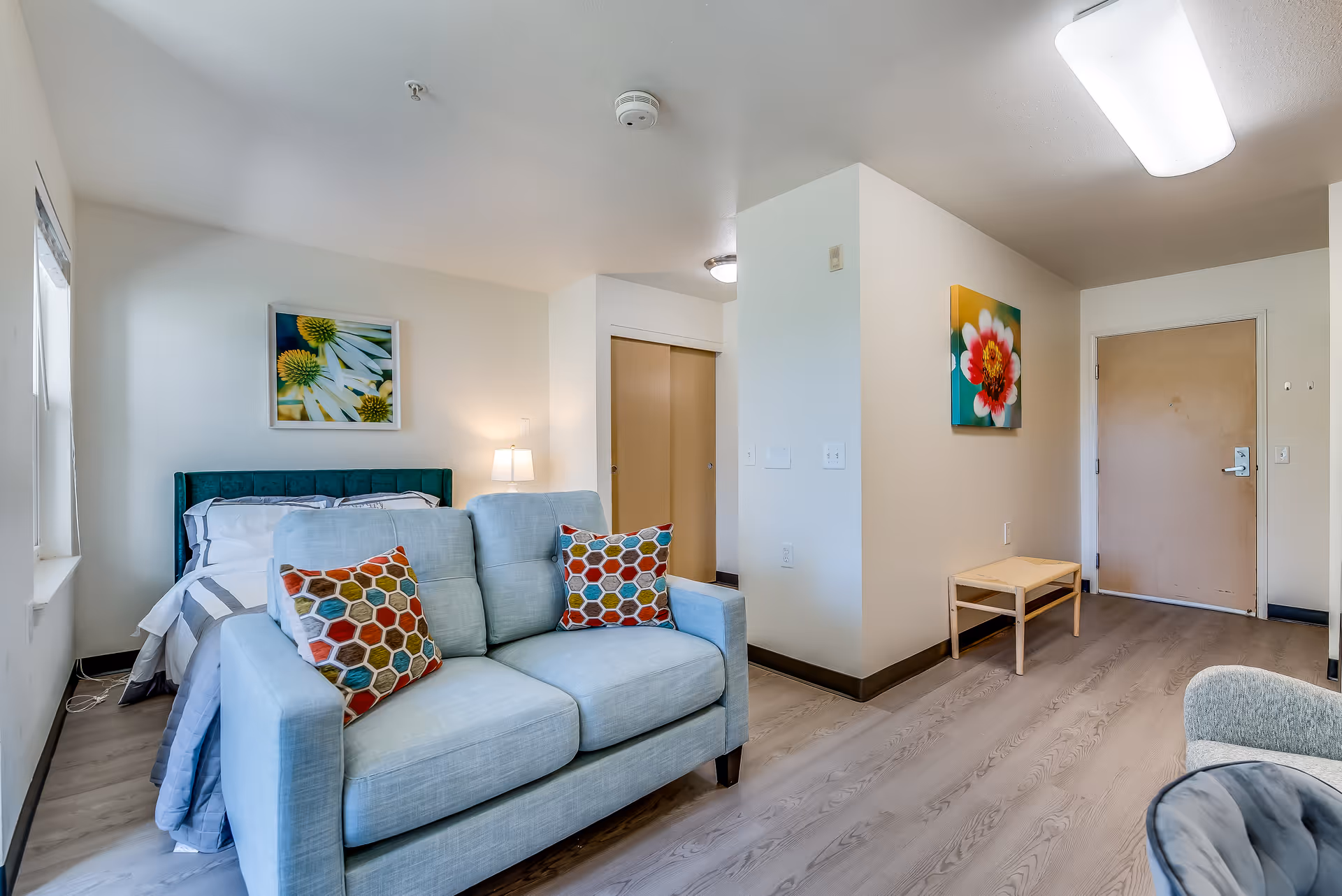 A bright and clean senior living studio apartment featuring a bed with white and gray bedding, a teal upholstered headboard, and a light blue sofa with two colorful hexagonal patterned pillows. The room has light wood flooring, white walls, a small wooden bench near the entrance door, and two floral paintings on the walls. A window on the left side allows natural light to enter the space.