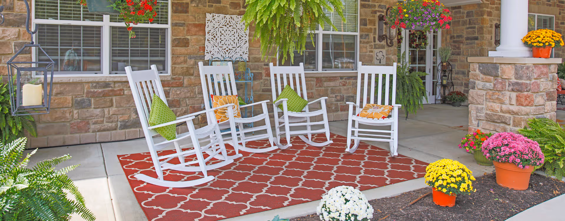 A cozy outdoor porch area with four white rocking chairs, each adorned with colorful cushions in green and orange patterns. The porch has a red patterned rug underneath the chairs and is surrounded by various potted flowers and green plants. The background features a stone wall with windows and a door leading inside the building.