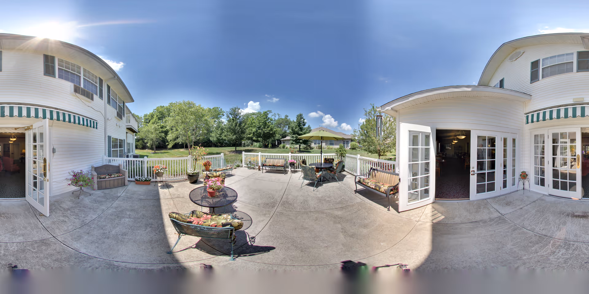 Outdoor patio area at Park Square Manor with concrete flooring, several benches and chairs with cushions, round tables with flower pots, a green umbrella, and white railings. The patio is adjacent to a white building with multiple glass doors and windows, and there is a grassy area with trees in the background under a clear blue sky.