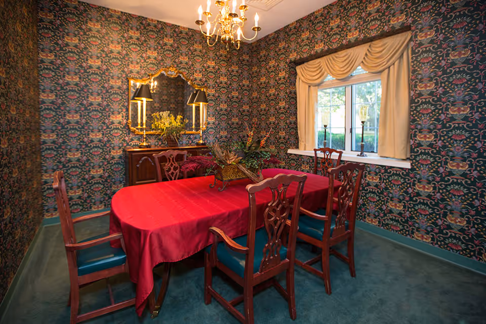 A dining room with a red tablecloth on an oval wooden table surrounded by six wooden chairs with green cushions. The walls are covered in a dark floral patterned wallpaper. A window with cream-colored curtains lets in natural light. A wooden sideboard with a decorative mirror and two lamps is against one wall, and a chandelier hangs from the ceiling.