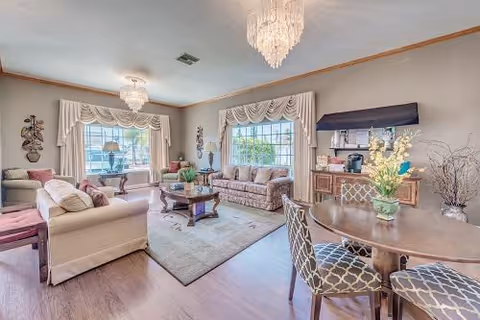 A bright and spacious living room with large windows covered by decorative curtains, two chandeliers, a beige sofa, a patterned sofa, a wooden coffee table on a rug, a round dining table with four patterned chairs, and a sideboard with a coffee maker and floral arrangement.