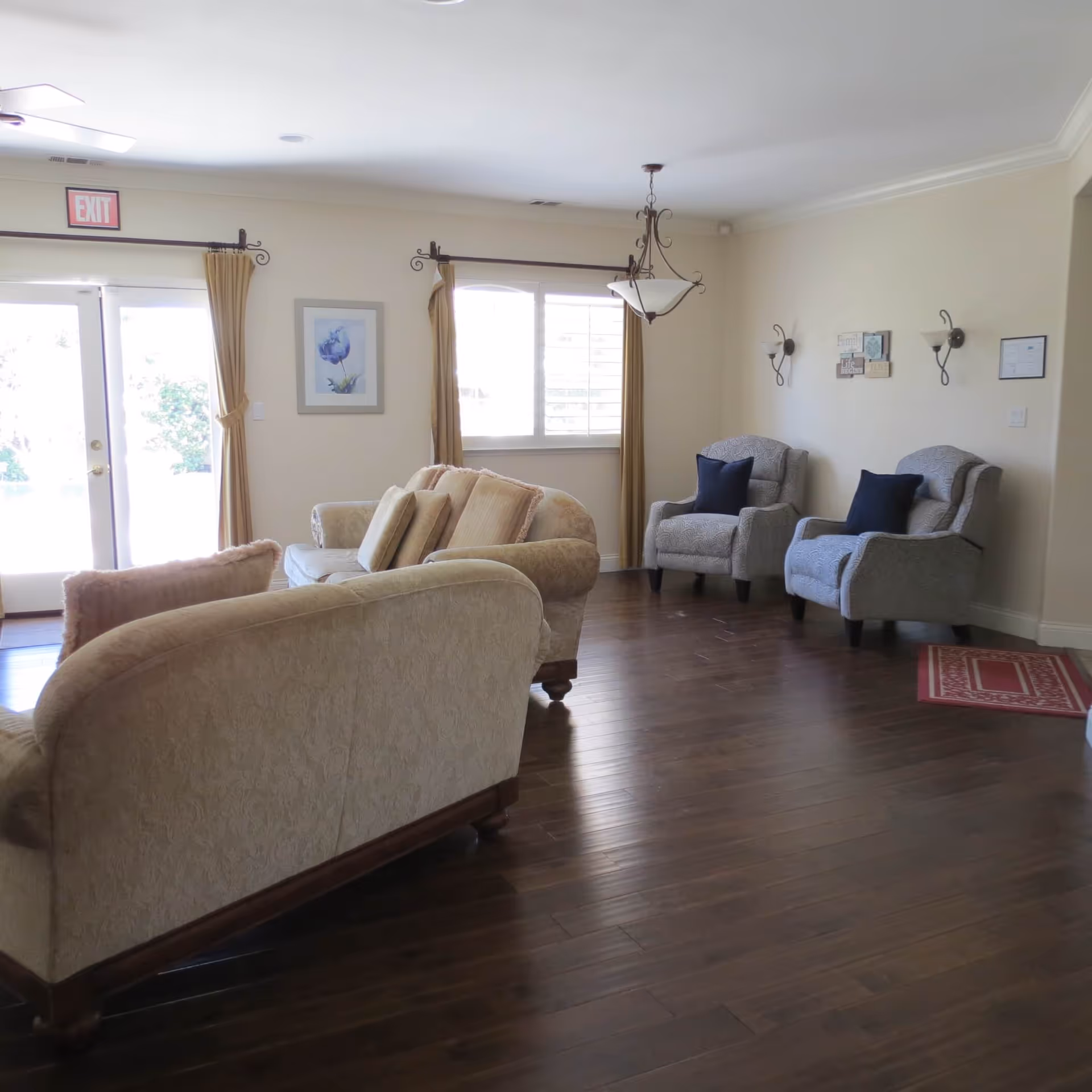 A bright living room with wooden flooring featuring two beige upholstered sofas with cushions, two gray armchairs with dark blue pillows, a window with beige curtains, a hanging light fixture, wall sconces, and a framed picture on the wall. There is an exit door with an exit sign above it.