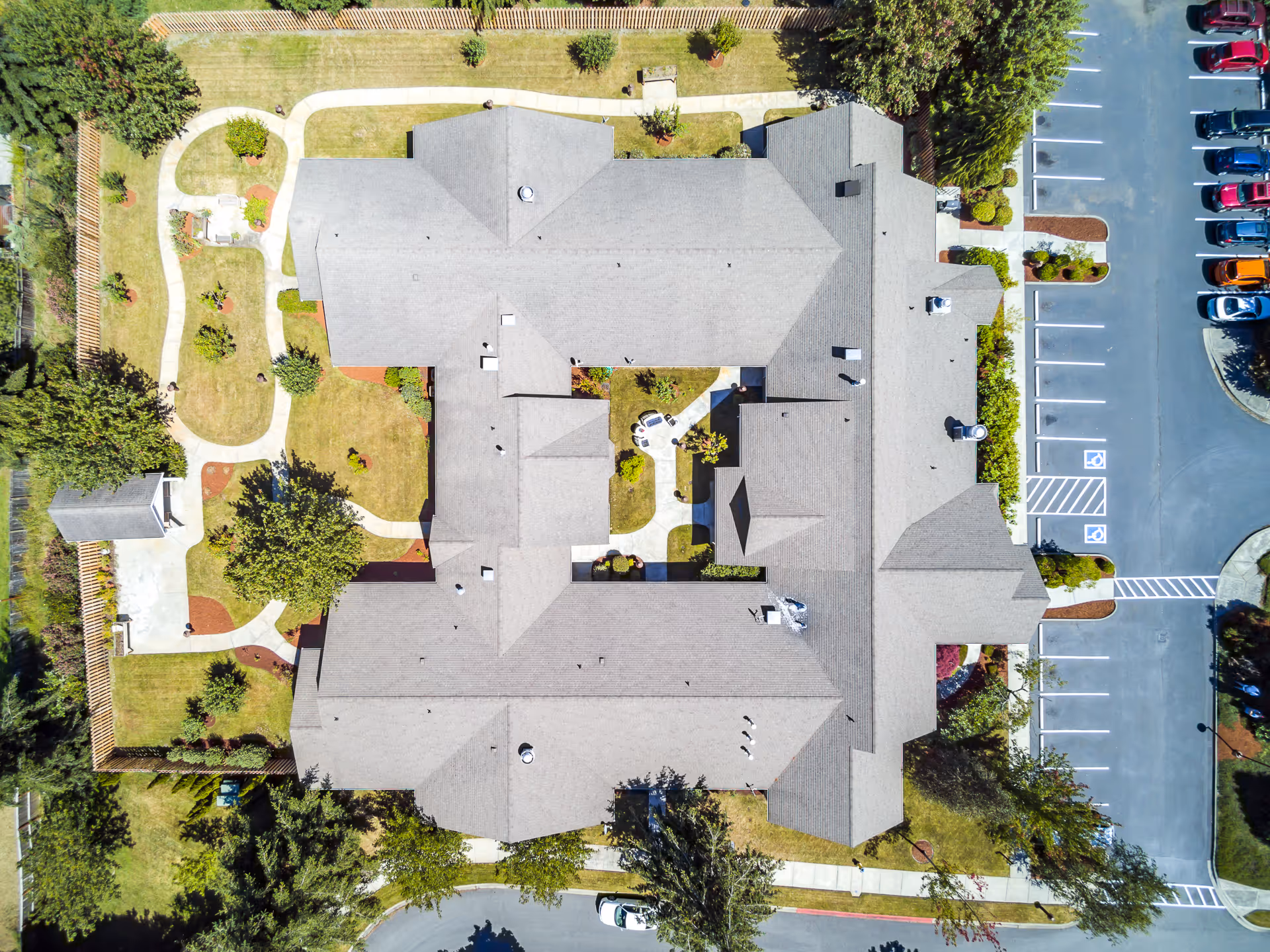 Aerial view of San Juan Villa Memory Care facility showing the building's roof, surrounding landscaped garden with walking paths, trees, and a parking lot with several parked cars.