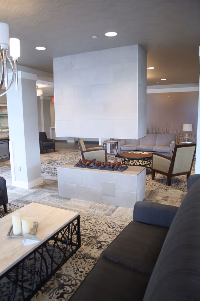 A modern living room area with a central rectangular electric fireplace surrounded by armchairs and sofas. The room features patterned carpeting, a wooden coffee table with candles, and soft lighting from ceiling fixtures and a table lamp in the background.