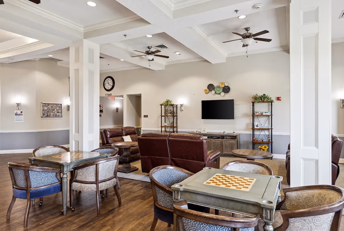 A spacious senior living common area with wooden floors, multiple tables and chairs, two ceiling fans, a wall-mounted TV, and leather sofas arranged around a coffee table. Shelves with decorative items and plants are placed against the walls, and a clock is visible above a hallway entrance.
