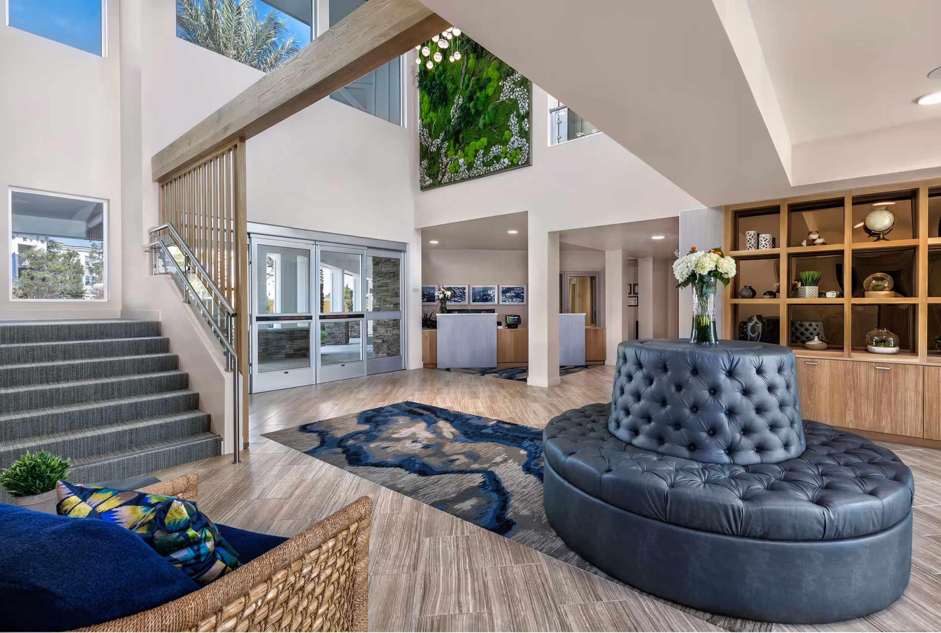 Bright and spacious senior living facility lobby with high ceilings, a modern circular tufted leather seating area with a vase of white flowers on top, a patterned blue and gray rug, a staircase with carpeted steps and metal railing, large windows letting in natural light, a green vertical garden on the wall, and a reception desk in the background.