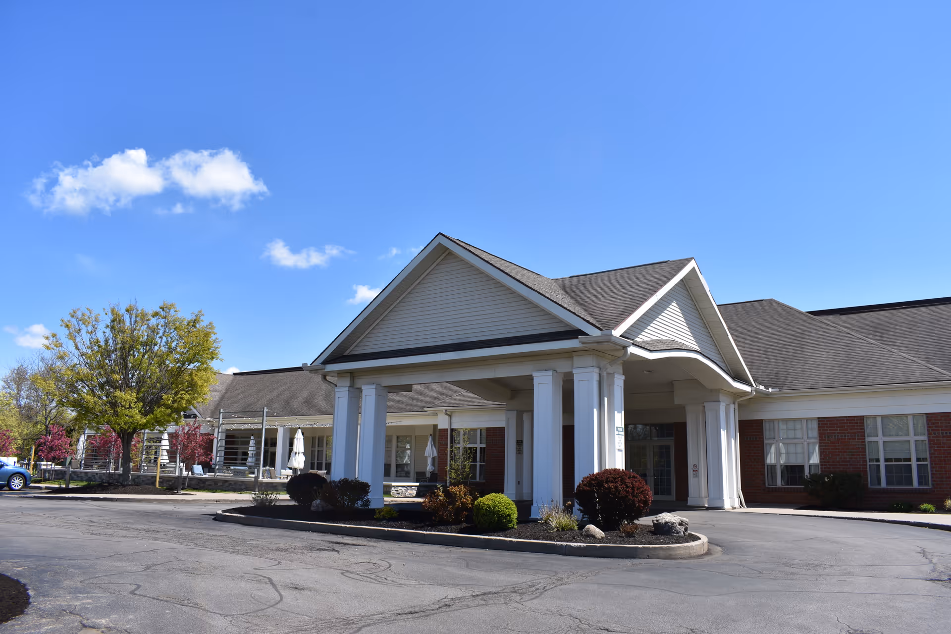 Exterior view of St. John's Meadows senior living facility showing the main entrance with white columns, a covered driveway, and a brick building under a clear blue sky with some trees and shrubs around.