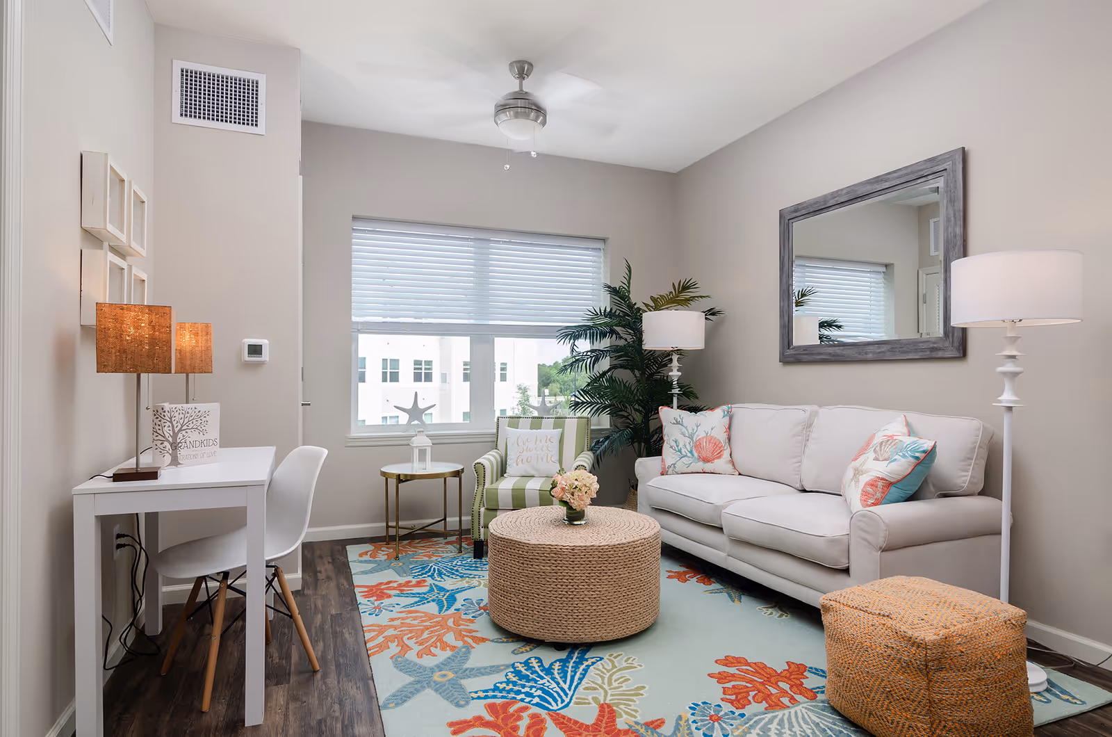 A cozy living room with a white sofa adorned with decorative pillows featuring coral and seashell designs. There is a large mirror on the wall above the sofa, a white floor lamp to the right, and a woven ottoman in front. A colorful rug with starfish and coral patterns covers the floor. To the left, there is a small white desk with a white chair and a table lamp with a textured shade. A green striped armchair with a decorative pillow sits near the window, next to a small round side table with a white lantern. A ceiling fan with a light fixture is mounted on the ceiling, and a large potted plant adds greenery to the corner.