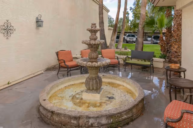 A tiered stone fountain in a courtyard patio surrounded by outdoor chairs and small tables with palm trees and parked cars visible in the background.